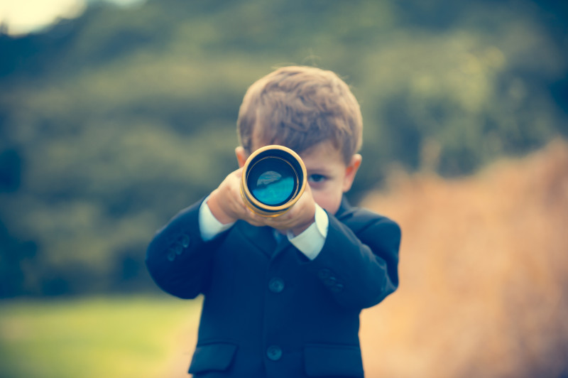 Young boy in a business suit with telescope. Small child wearing a full suit and holding a telescope. He is holding the telescope up to his eye. Business forecasting, innovation, leadership and planning concept. Shot outdoors with trees and grass in the background